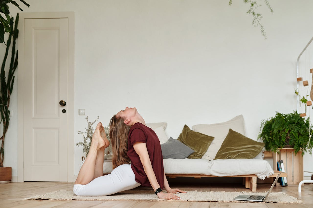 Woman stretching on a yoga mat indoors with a laptop nearby, embracing a healthy lifestyle.